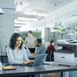 Xerox® C325 Colour Multifunction Printer in an office situation Professional woman typing on laptop at desk in office environment next to a Xerox® C325 Printer