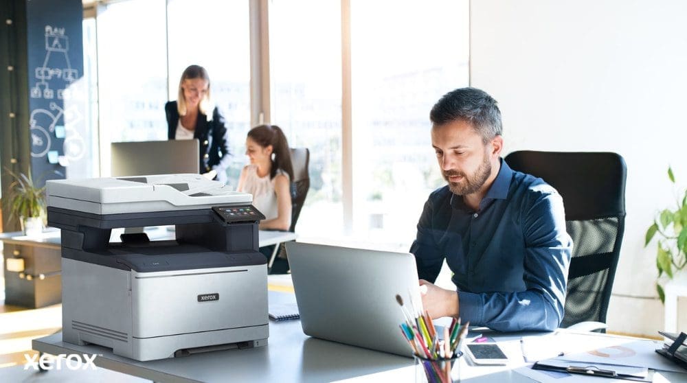 Xerox® Smart Start: Designed for Modern Workspace A person is seated at a desk, looking at a laptop with a Xerox C325 multifunction printer beside it. In the background, two other people are also working at their desks in a bright office environment.