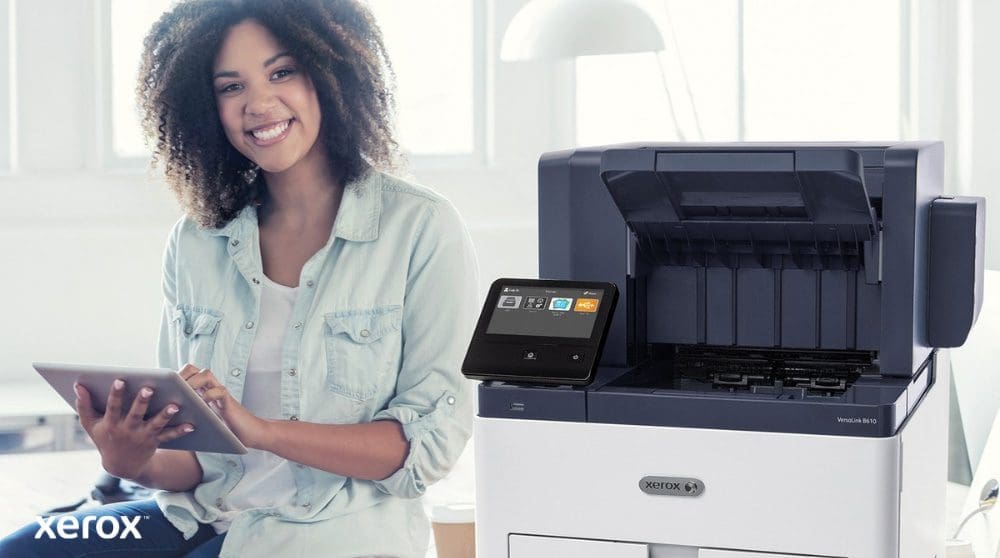 Xerox® Workplace Solutions: User-Friendly Integration A smiling person is seated next to a white Xerox VersaLink B610 printer, holding a tablet and looking at the camera, highlighting the user-friendly nature of Xerox solutions.